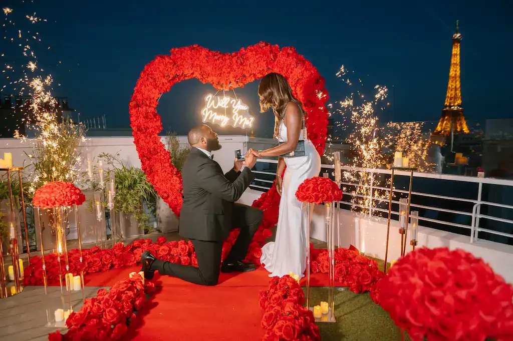 Man proposing to woman with Eiffel Tower in background, decorated with red roses and heart-shaped ar.